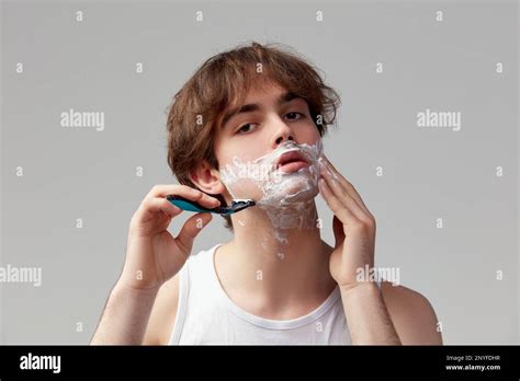 Portrait of young man shaving face with razor and cream, posing against grey studio background ...