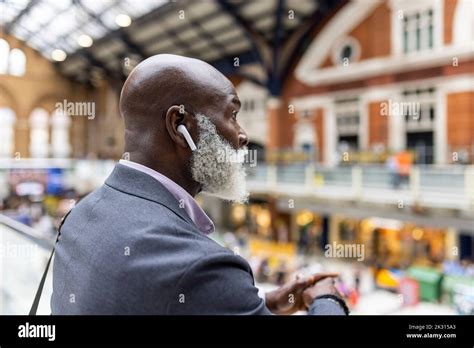 Bald senior commuter listening to music through wireless in-ear headphones at station Stock ...